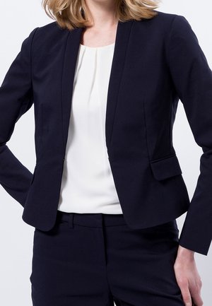 Femme portant un blazer bleu marine et un pantalon avec un chemisier blanc, debout avec une main sur la hanche devant un fond blanc.