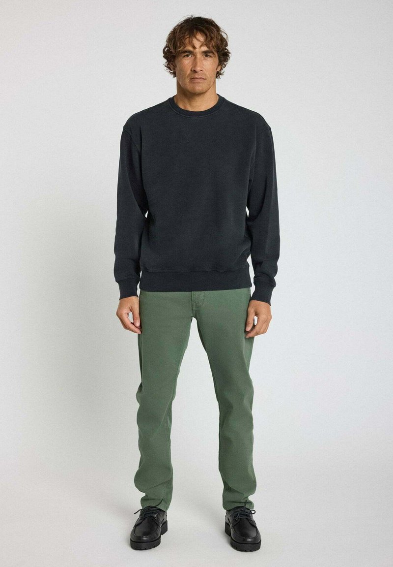 Sweatshirt noir à col rond avec des poignets côtelés, associé à un pantalon droit vert. Modèle portant des chaussures à lacets noires, se tenant sur un fond neutre.