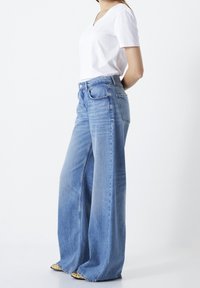 Mujer vistiendo unos pantalones de mezclilla de pierna ancha en azul claro, una blusa blanca de manga corta con escote en V y zapatos dorados de punta, de pie con las manos detrás de la espalda.