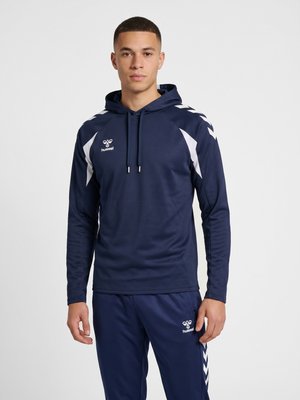 Młody mężczyzna ubrany w granatową sportową bluzę z kapturem i dopasowane spodnie z białymi wzorami typu chevron oraz logo Hummel, stojący na jednolitym tle.
