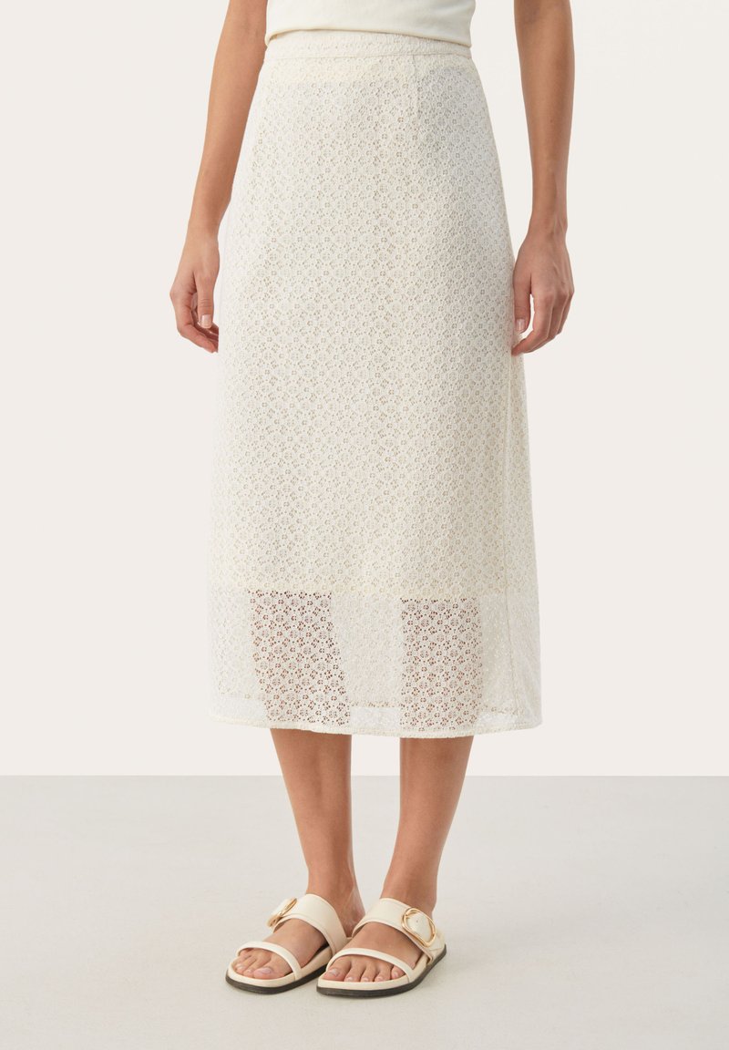 Jupe en dentelle crème avec un motif floral et une finition texturée ; longueur genou avec un ourlet transparent. Associée à des sandales beiges avec une boucle dorée.