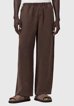 Mężczyzna ubrany w luźne brązowe spodnie z elastycznym pasem i brązowe sandały z odkrytymi palcami, stojący na jednolitym tle.