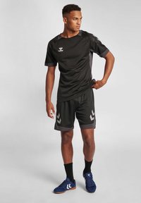 Zwarte sportshirt met gaasaccenten, gecombineerd met bijpassende shorts met grijze chevronpatronen. Schoeisel is blauw met een bruine zool.