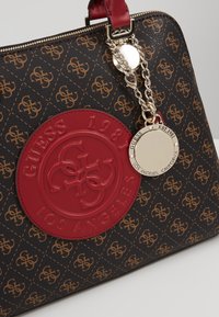 Sac à main marron à motifs avec des logos couleur fauve, un grand écusson rond rouge indiquant Guess 1981 Los Angeles, et une chaîne dorée avec une étiquette ronde Guess.