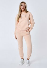 Perzikkleurige hoodie en jogger set, gemaakt van zachte stof, met een voorkantzak, koordjes en elastische boorden. Sneakers voegen contrast toe.