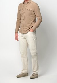 Beżowa koszula z długim rękawem z dwoma kieszeniami na piersiach, zestawiona z białymi spodniami i jasnoszarymi trampekami. Miękka tkanina o prostym wzorze.