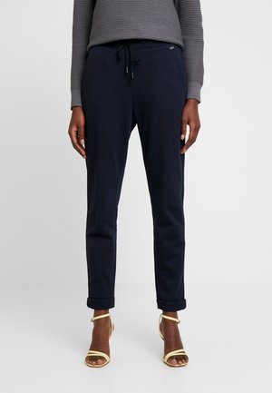 Femme portant un pantalon bleu marine foncé roulé, un haut gris texturé à manches longues et des sandales à talons hauts jaune clair à lanières, debout sur un sol blanc.