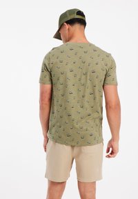 Alyvuogių žalia t-shirt su mažais mėlynais ir ruda raštais, dėvima su šviesiai smėlio spalvos šortais, demonstruojanti prigludusį dizainą ir trumpas rankoves.