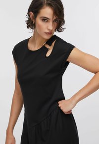 Robe noire avec manches courtes, encolure ronde avec une découpe, et un ourlet volanté. Fabriquée en tissu lisse et extensible avec un léger brillant.