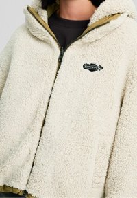 Cremefarvet oversized fleecejakke med struktureret stof, lynlås og hætte. Har en tan-farvet foring og et sort logo-lap.