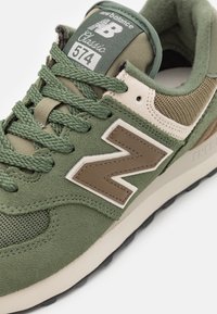 Oliwkowe sneakersy z zamszu i siateczki, z wyraźnym beżowym logo "N" i teksturowanymi sznurówkami, gumową podeszwą oraz wyściełanym kołnierzem.