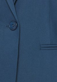 Tejido de blazer azul con una textura suave, que presenta un cierre de botón único y un detalle de bolsillo lateral. Diseño simple y color sólido.