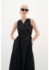 Schwarzes ärmelloses Kleid mit Wickeldesign, gerafftem Taillenbereich und Taschen. Glatter, leichter Stoff mit minimalistischem Schnitt.