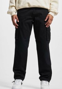 Pantalons cargo noirs avec poches latérales, fabriqués en tissu durable. Coupe décontractée avec une jambe droite. Portés avec des baskets blanches et un sweat à capuche beige.