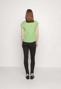 Camisa verde de manga curta com riscas verticais combinada com calças pretas ajustadas e sapatos pretos; vista de costas num fundo simples.