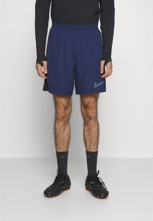 Mężczyzna w granatowych sportowych spodenkach z logo Nike, czarnej bluzie z długim rękawem, czarnych skarpetkach Nike oraz czarnych korkach z pomarańczowymi kołkami.