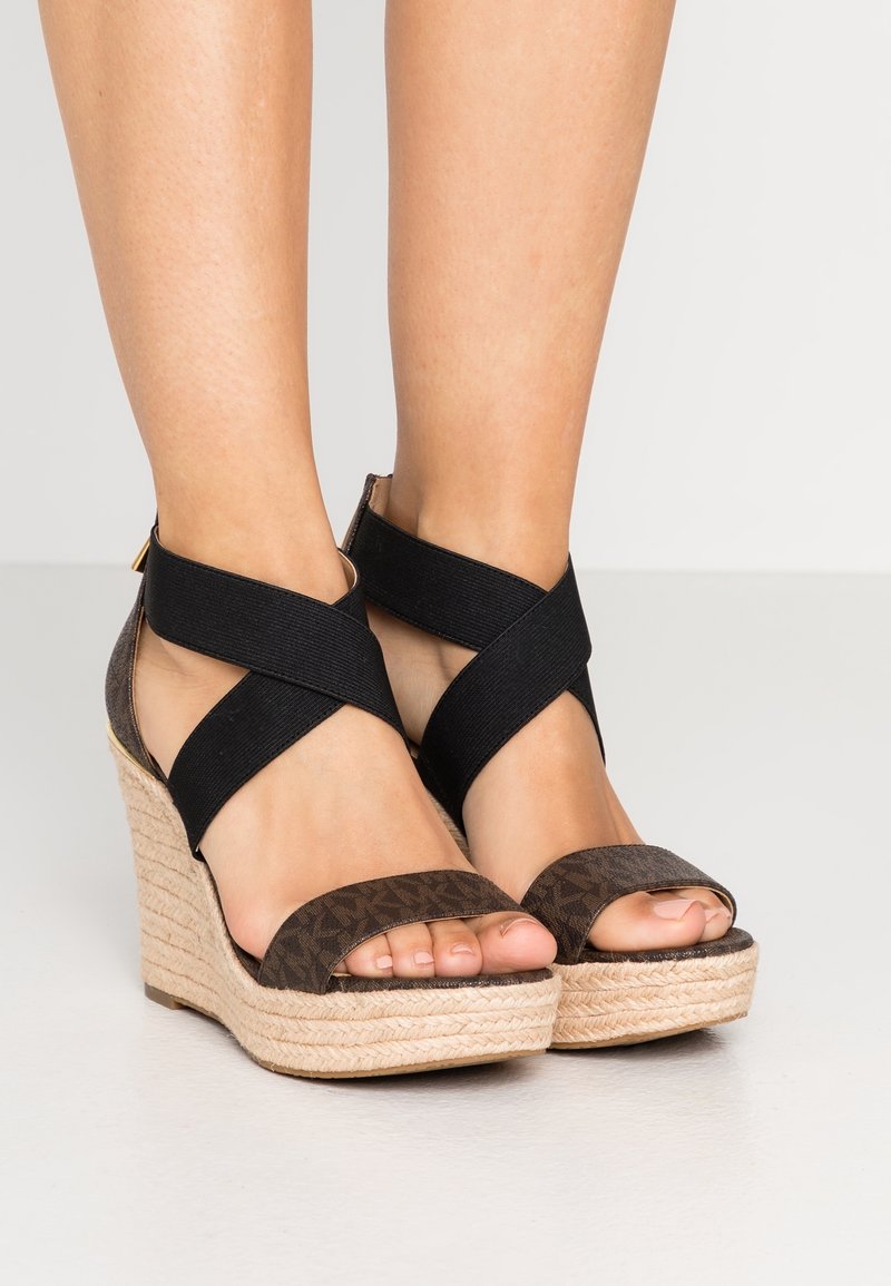 Pieds portant des sandales compensées marron et noires avec des brides élastiques croisées et des semelles plateformes tissées sur une surface blanche.