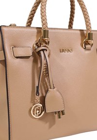 Beige læderhåndtaske med flettede håndtag, guldfarvet beslag og vedhæng med et logo. Har en tekstureret overflade og struktureret design.