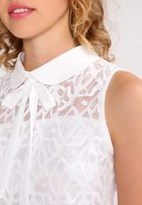 White sleeveless blouse with a textured lace design, a soft collar, and a bow tie detail at the neckline.
