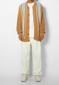 Brown knitted cardigan with blue and cream vertical stripes, featuring brown buttons, worn over a white t-shirt and cream pants.