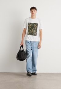 Ein weißes T-Shirt mit einem strukturierten, grün-gold gemusterten Quadrat, kombiniert mit hellblauen, lockeren Jeans und einer schwarzen Tasche. Schwarze Schuhe.