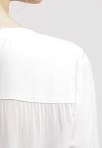 Vit blus med en slät textur, rundad halsringning och lätt veckad bak. Lätt tyg med en subtil lyster.