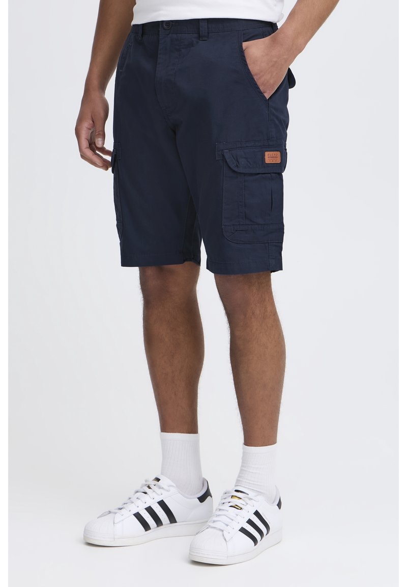 Shorts cargo bleu marine en coton avec deux poches latérales et une étiquette en patch, associés à des baskets blanches avec des rayures noires et des chaussettes côtelées.