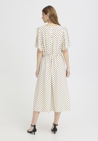 Femme portant une robe midi crème à pois noirs, manches volantées, taille cintrée, et talons à bride noire à la cheville, vue de dos.