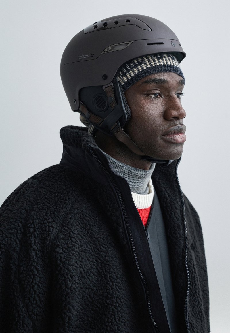 Dark helmet with ventilation holes, worn over a knitted striped beanie. Model is dressed in a black sherpa coat and layered, muted-tone clothing.