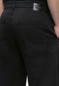 Pantalon noir avec deux poches arrière et un patch en cuir de la marque "DRYKORN" sur la ceinture, porté par une personne avec la main dans la poche latérale.