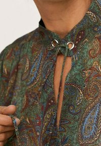 Blouse à motif cachemire en vert, bleu et marron avec un col montant, des éléments métalliques argentés, et une fermeture à nouer au niveau de l'encolure.
