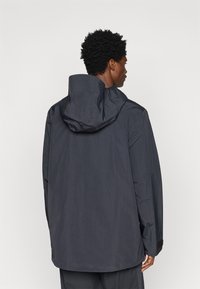 Muži z pohľadu zozadu, ktorý má na sebe tmavosivú kapucňovú bundu s dlhými rukávmi na neutrálnom bielom pozadí.