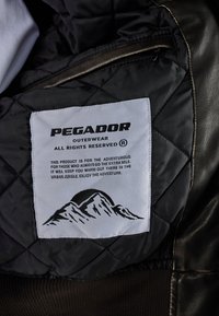 Fekete steppelt belső bélésű dzseki, amelyen egy hegyek grafikáját ábrázoló szövetcímke található, fehér háttérrel. A címkén a felirat: "Pegador Outerwear."