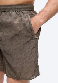 Mees, kes kannab oliivrohelisi Diesel lühikesi pükse elastse vöökohaga, parem käsi taskus, näidates riide kanga tekstuuri ja brändi mustrit.