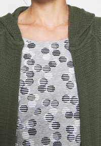 Olivově zelený pletený kardigan přes šedé tričko s vzorem černobílých pruhovaných kruhů. Měkká textura, volný střih.