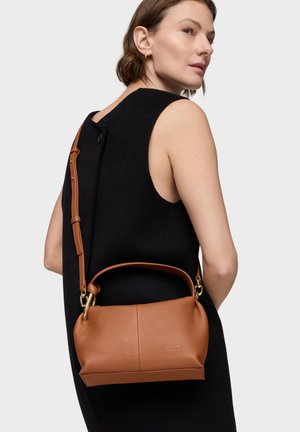 Mujer con vestido negro sin mangas llevando un bolso de cuero marrón medio con asa superior y correa de hombro ajustable sobre la espalda.