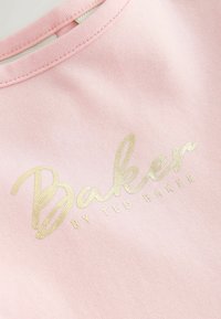 Pink stof i nærbillede med guldskrift, der lyder "Baker by Ted Baker" nær halsudskæringen.