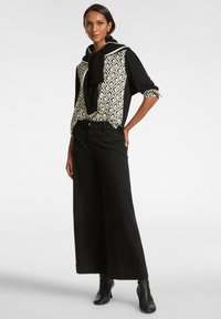 Pantalones de pierna ancha negros combinados con una blusa estampada que presenta un diseño de diamantes en crema y negro; estilizados con un suéter negro atado alrededor del cuello.