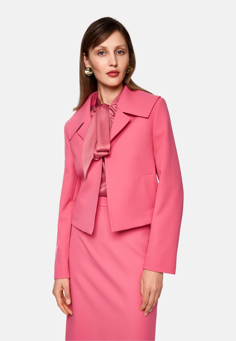 Rosa kurz geschnittene Jacke mit einem großen Kragen, kombiniert mit einer rosa Bluse, die über eine Bindegarnitur verfügt. Der Stoff wirkt glatt und strukturiert.