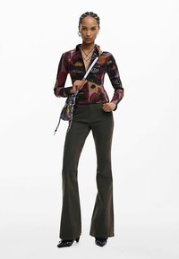 Mehrfarbiges Langarmshirt mit Grafikdesign, kombiniert mit olivgrünen Schlaghosen und glänzenden schwarzen Schuhen, accessoriert mit einer kleinen Tasche.