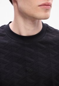 Jeune homme portant un t-shirt noir texturé à col rond avec un motif géométrique subtil et un petit logo « BOSS », visible du menton à la poitrine.