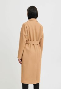 Manteau en mélange de laine beige avec une taille ceinturée, un col cranté et des manches longues. Le dos présente une couture centrale et un design minimaliste.