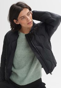 Zwart gebreid jack met een ritssluiting aan de voorkant en een geribbeld kraag, gecombineerd met een lichtgroene gebreide trui. Glad materiaal en slim fit ontwerp.