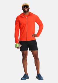 Feloranje lange mouwen zip-up hoodie, zwarte shorts, blauwe hardloopschoenen en een gele pet. Houdt een dumbbell en een waterfles vast.