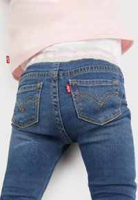 Blå denimjeans med gula sömmar, med en distinkt design på bakfickan. Bärs med en ljusrosa topp och grå midja.