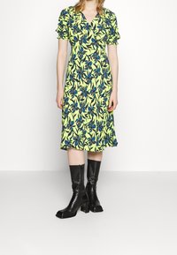 Donna che indossa un vestito midi giallo con motivo floreale blu e nero, maniche corte a palloncino, abbinato a stivali neri con tacco e al polpaccio su sfondo bianco.