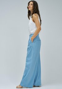 Un pantalon en denim bleu clair à jambes larges avec des poches latérales, taille haute, et une texture lisse, associé à un top sans manches blanc avec des motifs floraux discrets.