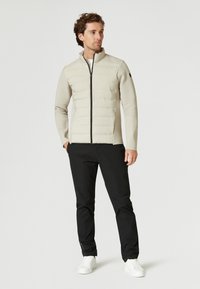 Ljusbeige pufferjacka med dragkedja fram och hög krage, kombinerad med svarta byxor och vita sneakers, som visar en modern och åtsittande design.