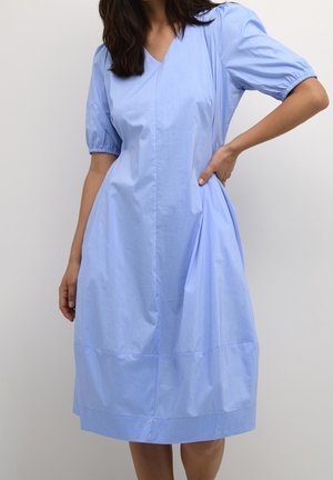 Femme portant une robe midi bleu clair à manches courtes bouffantes et col en V, debout avec une main sur la hanche.
