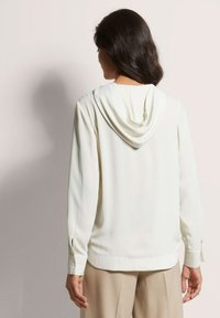 Witte hoodie blouse met lange mouwen, zachte stof en knoopmanchetten; gladde textuur en een iets losse pasvorm aan de onderkant.
